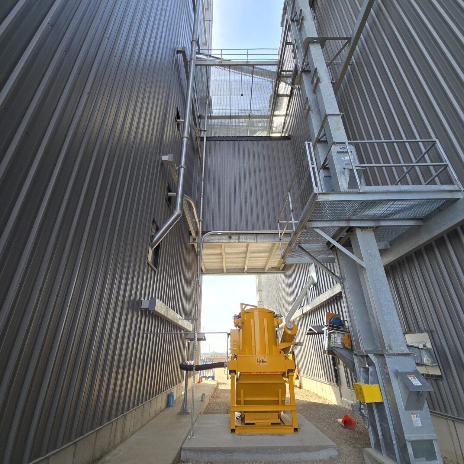 Yellow Hi‑Vac industrial vacuum unit centered between tall corrugated metal walls with overhead catwalk and steel ladder.