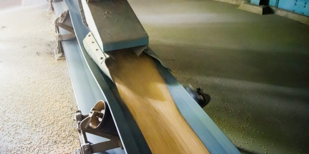 Blue conveyor chute feeding flowing grain into a hopper, industrial bulk handling at a Hi‑Vac facility